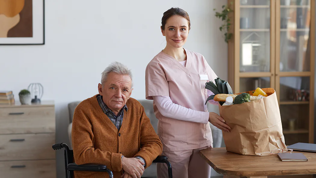 Alltagsbegleiterin bringt älterem Mann im Rollstuhl frische Lebensmittel nach Hause.