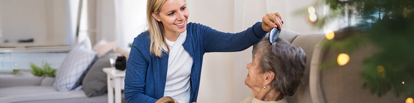 Alltagsbegleiterin kämmt liebevoll die Haare einer älteren Frau und hält dabei ihre Hand im Wohnzimmer.