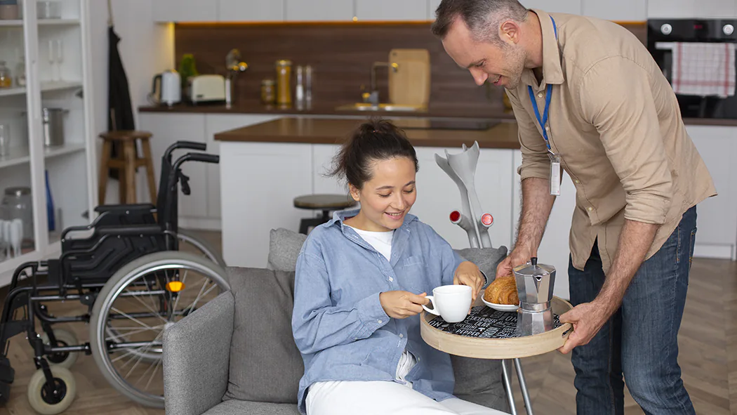 Betreuungskraft reicht pflegebedürftiger Frau Essen und Trinken.