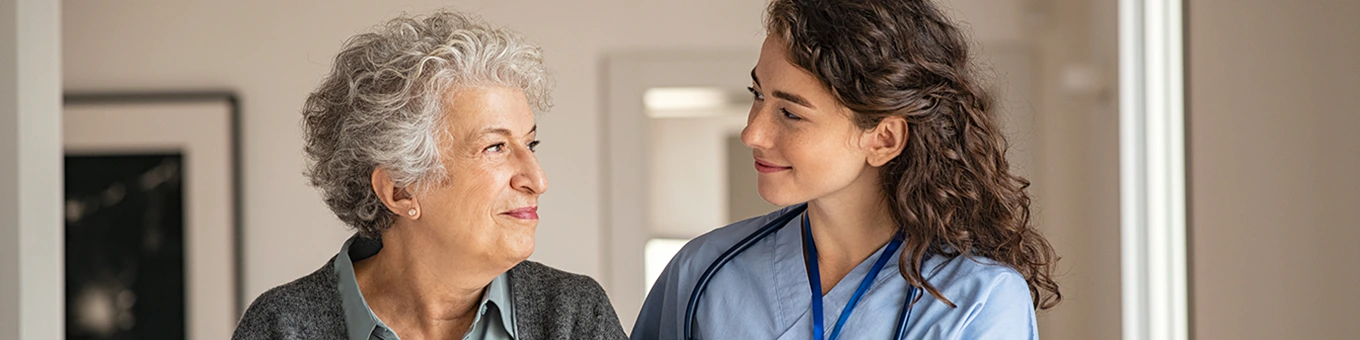 Pflegehilfskraft und Seniorin lächeln sich an.