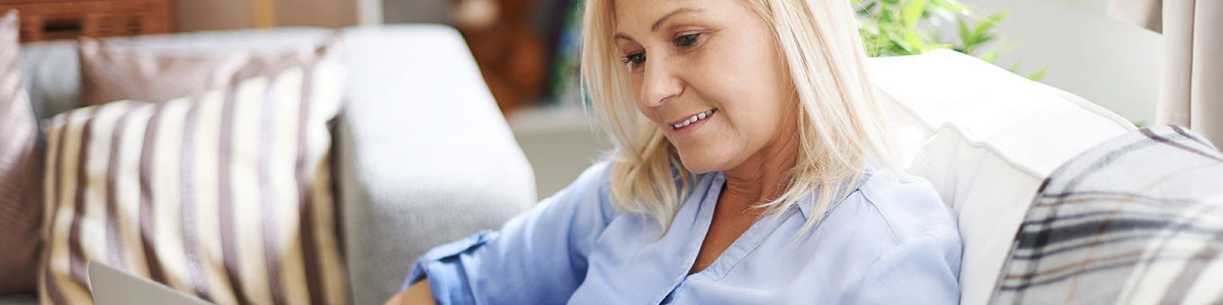 Frau mittleren Alters sitzt mit Laptop auf dem Schoss auf der Couch und lächelt.