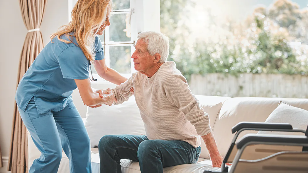 Betreuungskraft hilft pflegebedürftigen Mann von der Couch hoch.