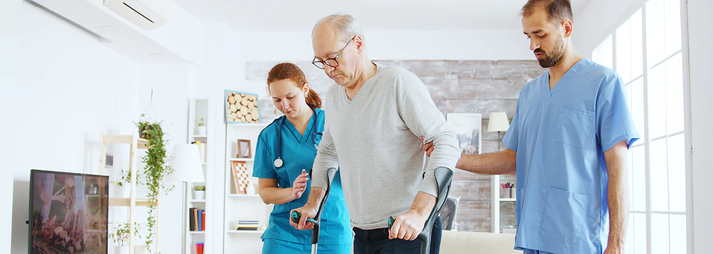Eine Pflegerin und ein Pfleger unterstützen einen älteren Patienten beim Laufen mit Krücken