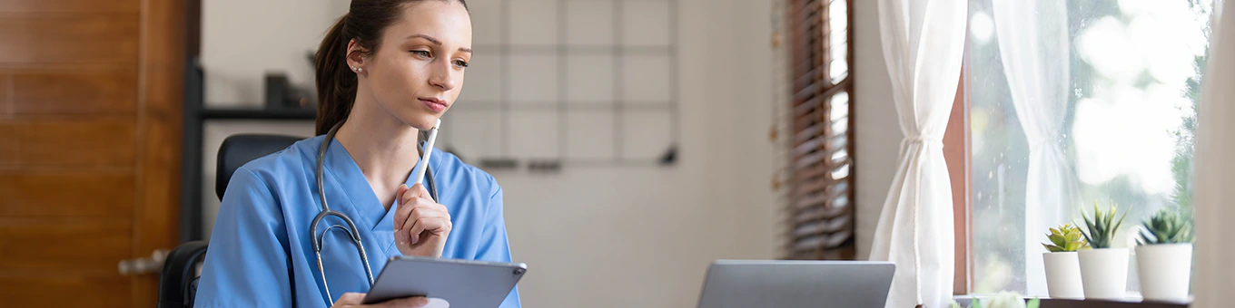 Pflegekraft sitzt am Schreibtisch, bildet sich konzentriert mit Tablet und Laptop weiter.