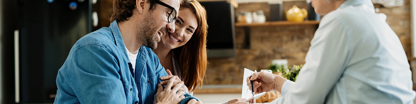 Ein lachendes Paar, bei dem der Mann im Rollstuhl sitzt, führt ein beratendes Gespräch mit einer Pflegeberaterin.